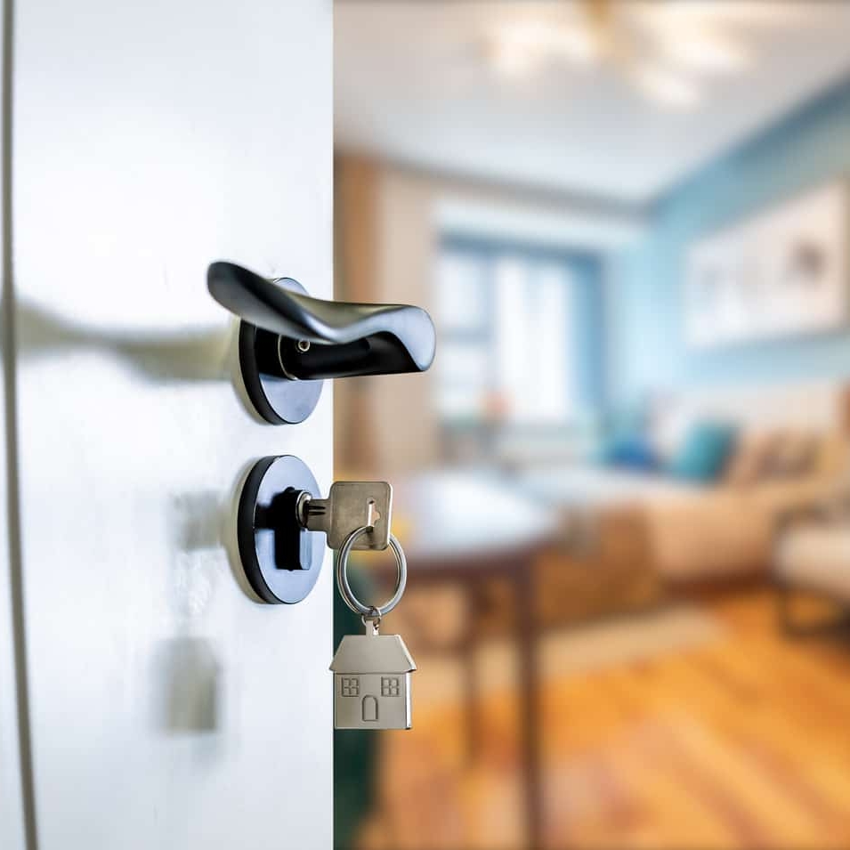 Close-up of an open door with a key in the lock, featuring a house-shaped keychain. A blurred living room is visible in the background, suggesting a perfect entry for estate agent websites to showcase property interiors.