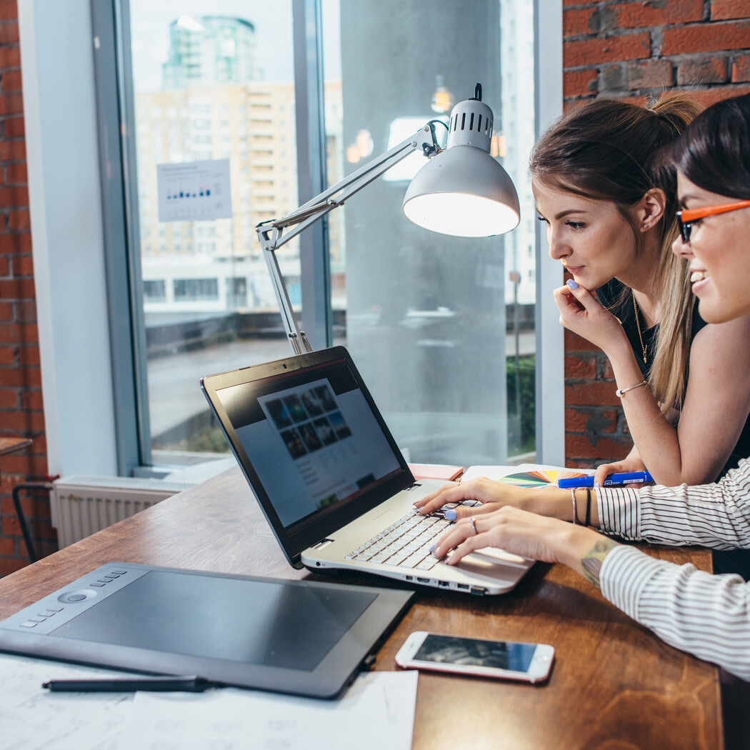Two women sit at a desk in a modern office, closely looking at a laptop screen while discussing how often to refresh your site, with papers, a tablet, and a phone spread out on the wooden table.