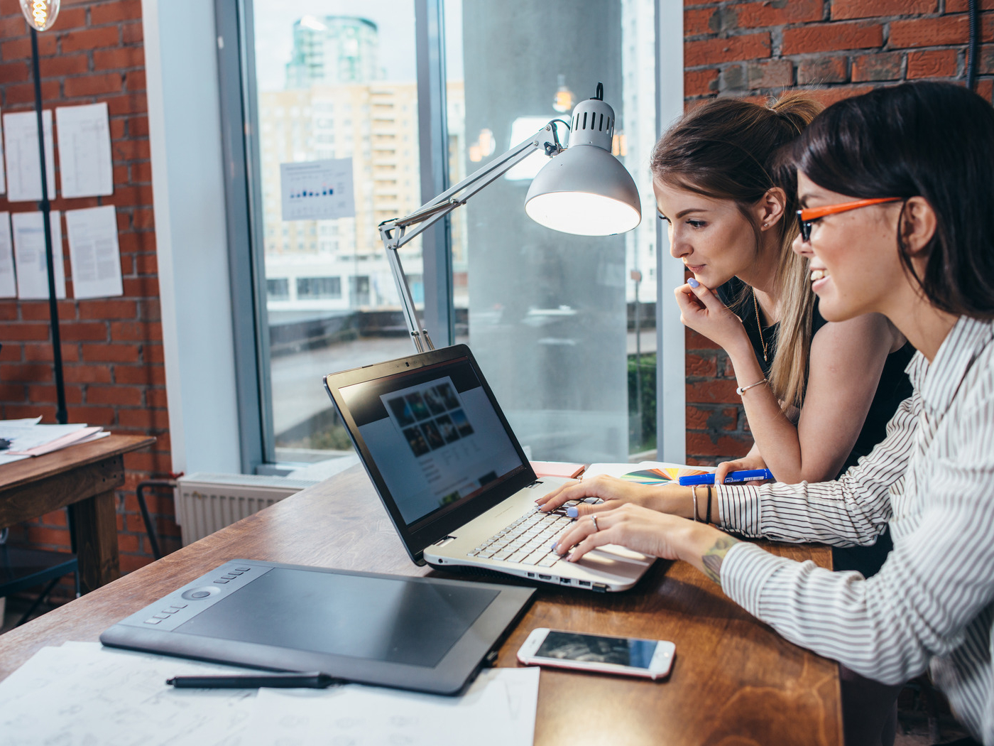 Two women sit at a desk in a modern office, closely looking at a laptop screen while discussing how often to refresh your site, with papers, a tablet, and a phone spread out on the wooden table.
