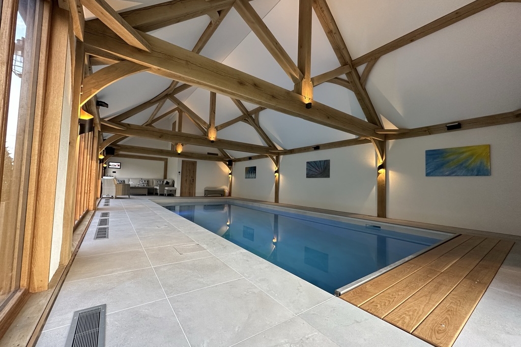 Indoor swimming pool with wooden beams on the ceiling, large windows on one side, a lounge area at the far end, and abstract artwork on the walls—designed with Cartlodge Company’s signature touch for stylish spaces.