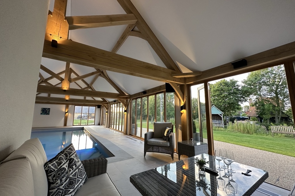 Indoor pool room with exposed wooden beams crafted by Cartlodge Company, glass doors opening to a garden, modern seating area, and a glass table set with wine glasses and pillows.