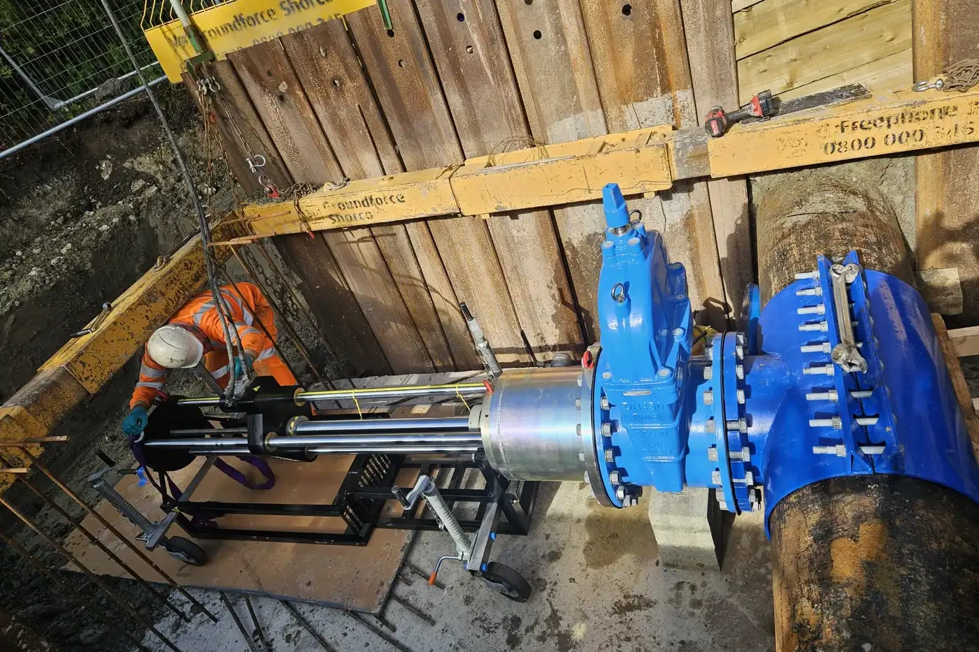 A worker operates machinery to install or maintain a large blue water valve on an underground pipeline at a construction site.