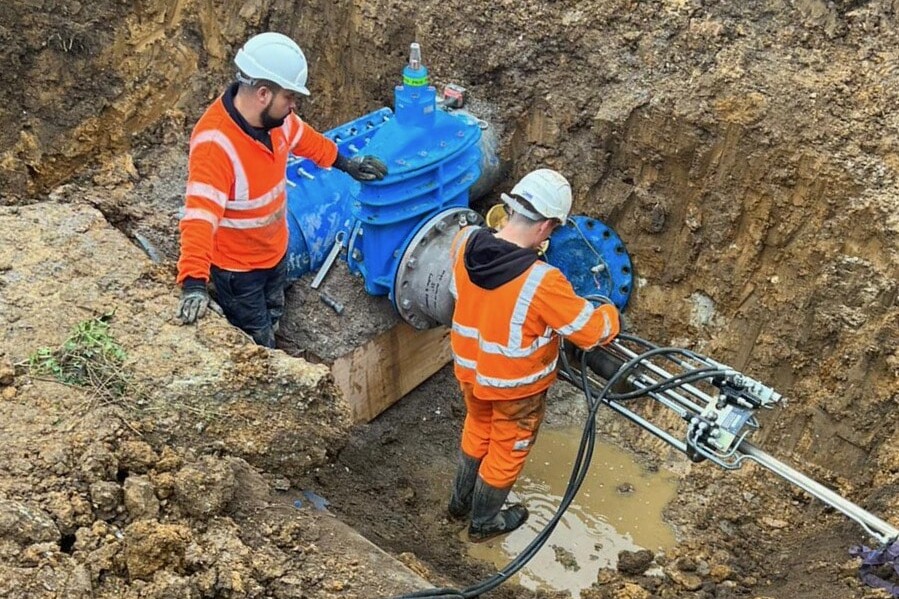 Two workers in orange safety gear and helmets install large blue water pipes and valves in a muddy trench.