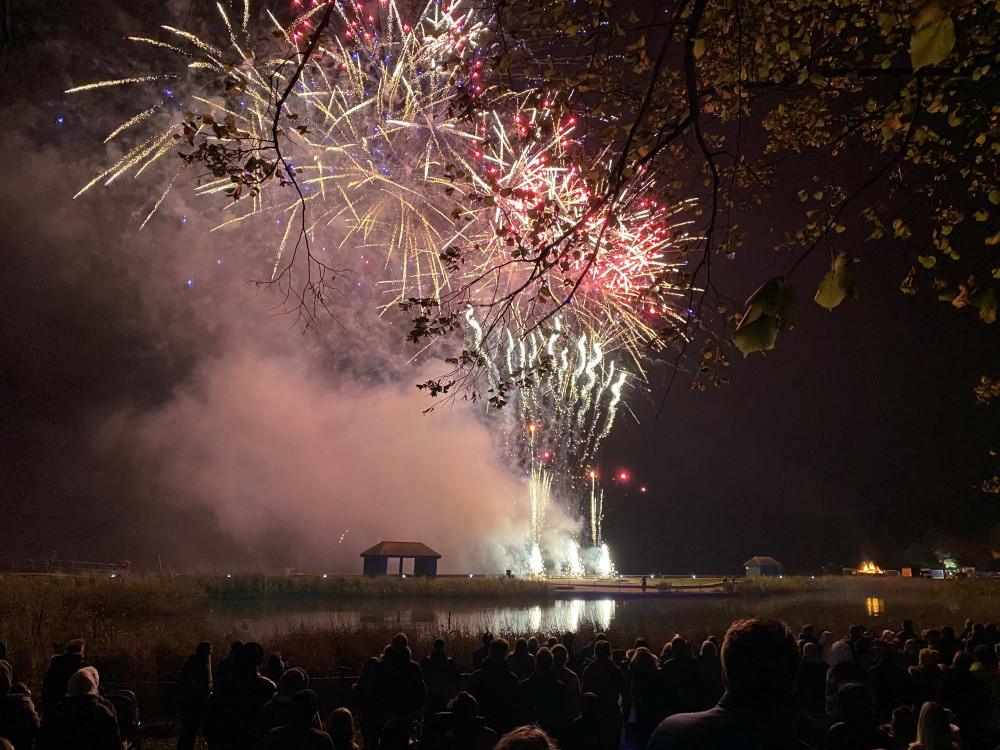 A crowd watches colorful fireworks light up the night sky above a lake at the Maldon Firework Display, with smoke rising and trees framing the annual spectacle.