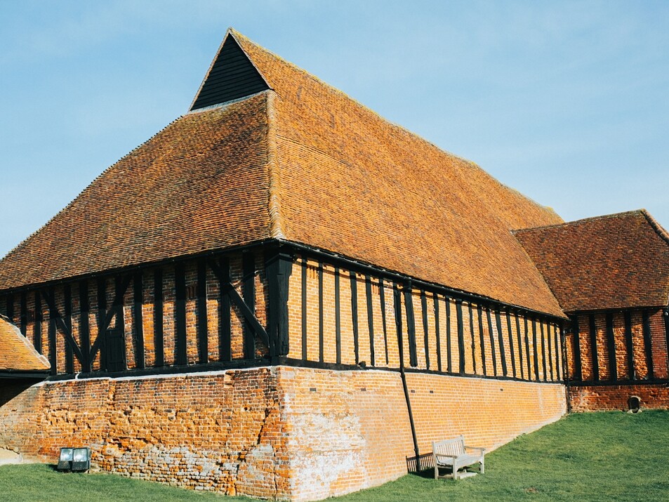 A large, timber-framed barn with steep, orange-tiled roof and brick lower walls at Cressing Temple Barns, set on a grassy area under a clear sky—an ideal venue for the 2025 Essex Christmas Fair.