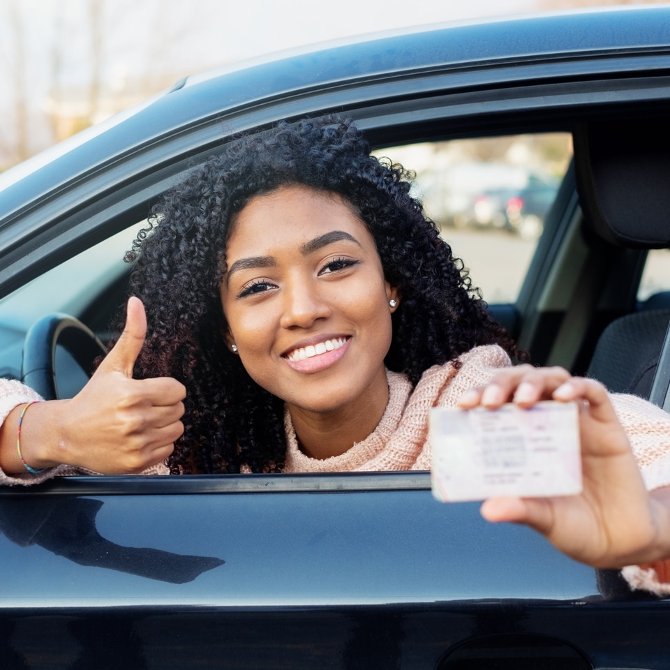 A person sitting in a car smiles, gives a thumbs up, and holds a driver's license out the window after completing driving lessons with Mutlows Driving School.