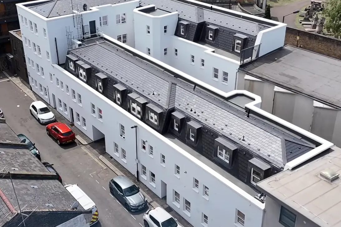 Aerial view of a modern, white apartment building with multiple floors and dormer windows, surrounded by parked cars on a narrow street—a glimpse behind the scenes (BTS) of urban Special Works in progress.