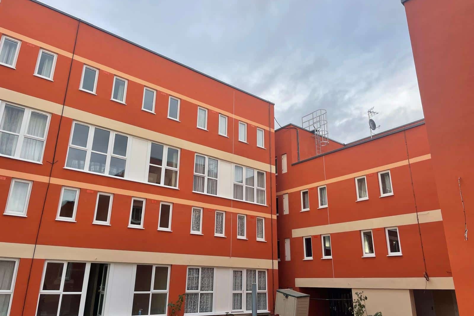 Red-orange apartment building with several white-trimmed windows, some open, and a small courtyard area with benches and plants under a cloudy sky—part of the Special Works BTS development.