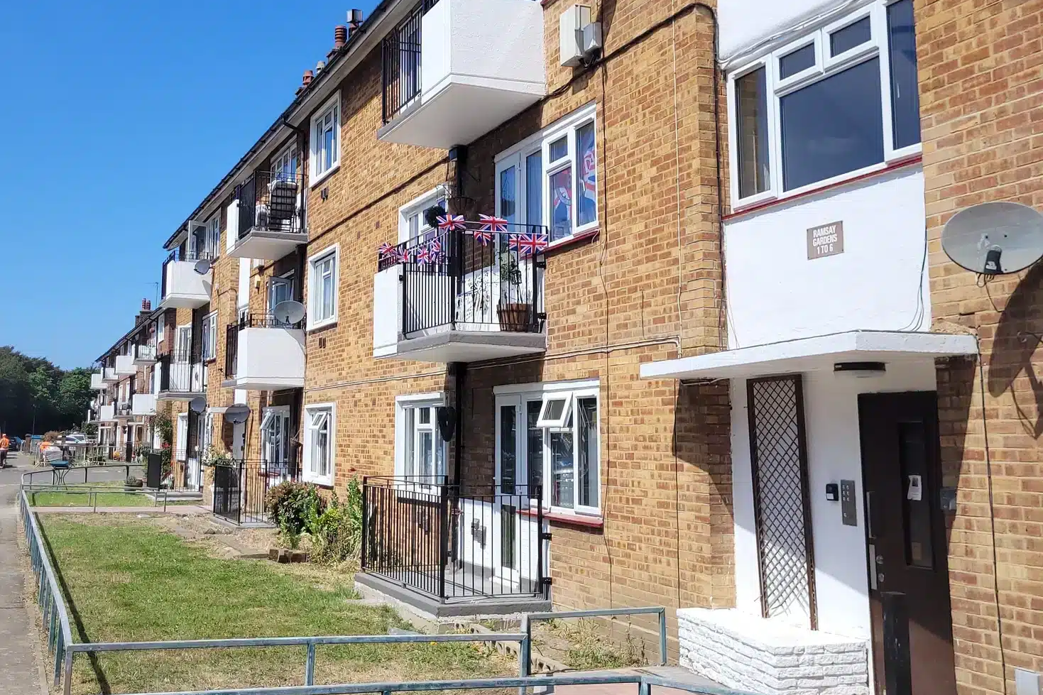 A row of brick apartment buildings with balconies, some decorated with Union Jack flags, and a small grassy area in front—perfect for BTS fans or those seeking a special works vibe in the heart of the city.