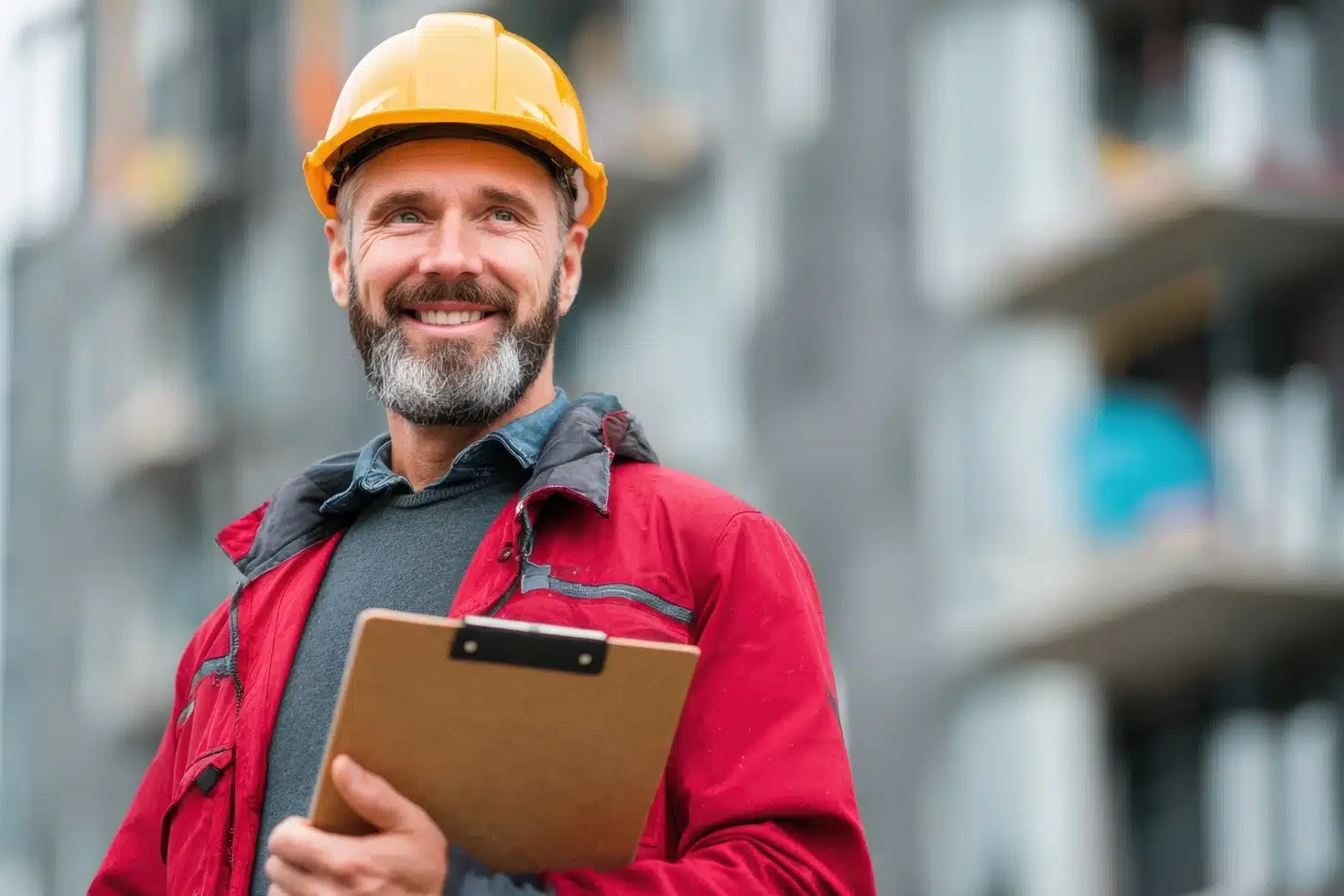 A man in a red jacket and yellow hard hat holds a clipboard and smiles, standing in front of a blurred building site, ready to serve as your Compliance Assistant.