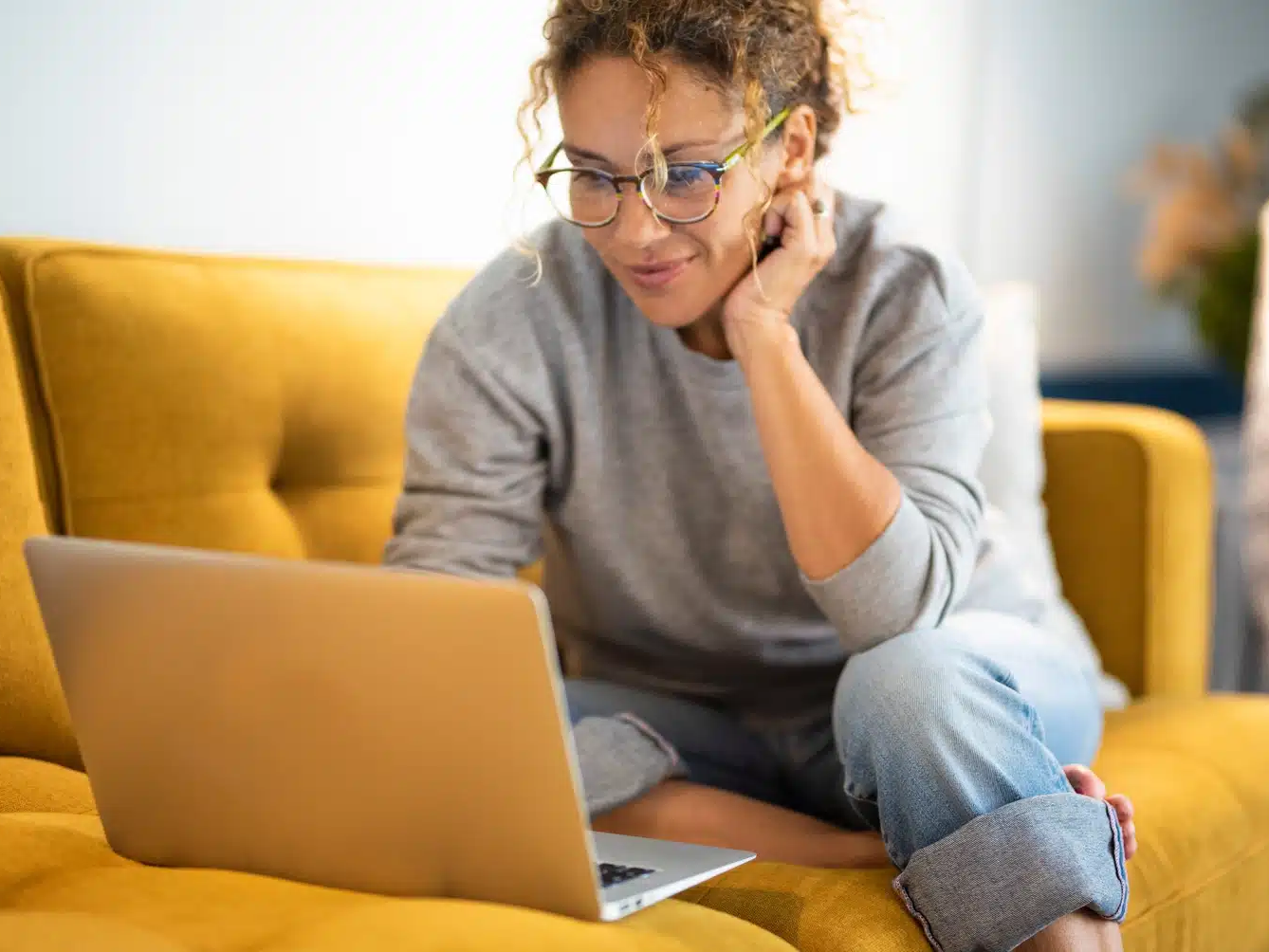 A person with curly hair and glasses sits on a yellow sofa, looking at a laptop screen with a slight smile, perhaps researching website development cost for 2026.